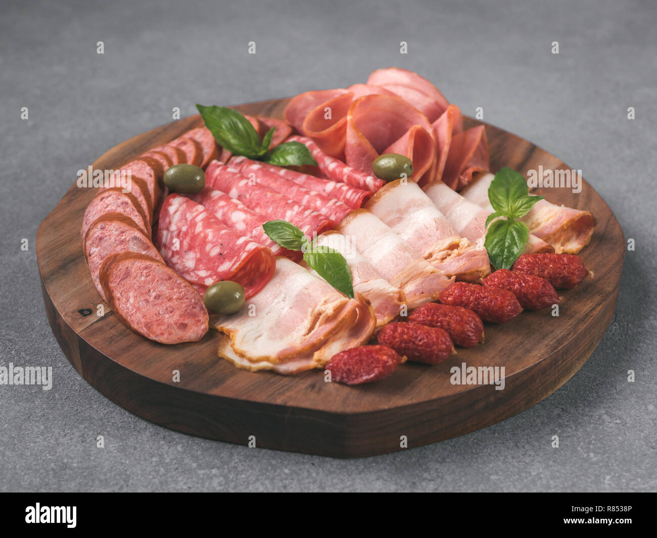 Antipasto set platter on wooden plate over gray stone. Cold smoked meat plate with sausage,sliced ham,prosciutto, bacon, olives and basil. Appetizer on wooden tray cut tree sawed imitation. Copy space Stock Photo