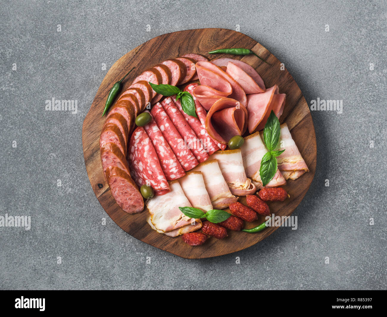 Antipasto set platter wooden plate on gray stone. Cold smoked meat plate with sausage, sliced ham,prosciutto,bacon,olives,basil. Appetizer on wooden tray cut tree sawed imitation. Copy space. Top view Stock Photo