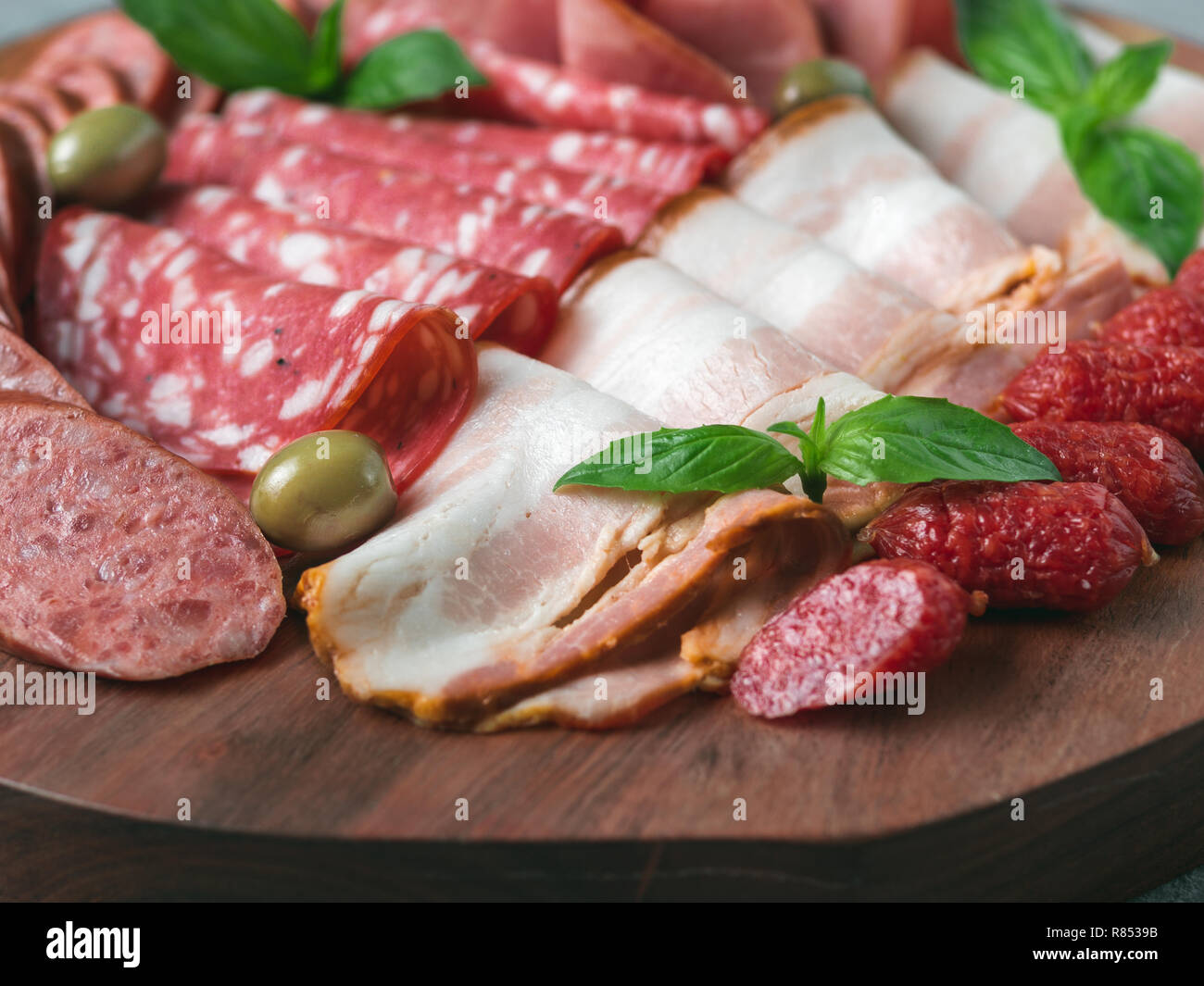 Antipasto set platter on wooden plate close up. Cold smoked meat plate with sausage,sliced ham,prosciutto, bacon, olives and basil. Appetizer on wooden tray cut tree sawed imitation. Copy space Stock Photo