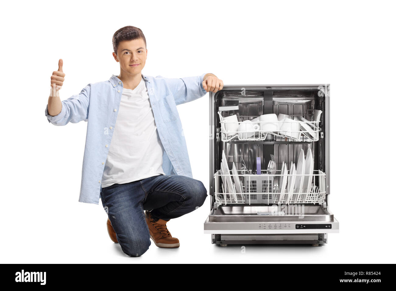 Young guy kneeling next to a loaded dishwasher with thumb up isolated on white background Stock Photo