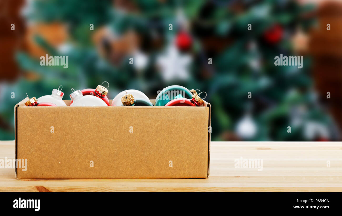 Cardboard box with colorful glass Christmas balls on wooden table against decorated Christmas tree on a blurred background. Copyspace. Stock Photo