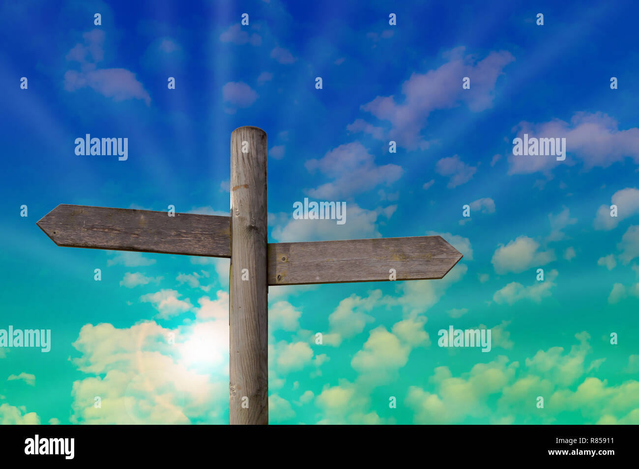 A wooden signpost with two empty arrows for your own text. Background with a light cloudy sky and light sun beams. Stock Photo
