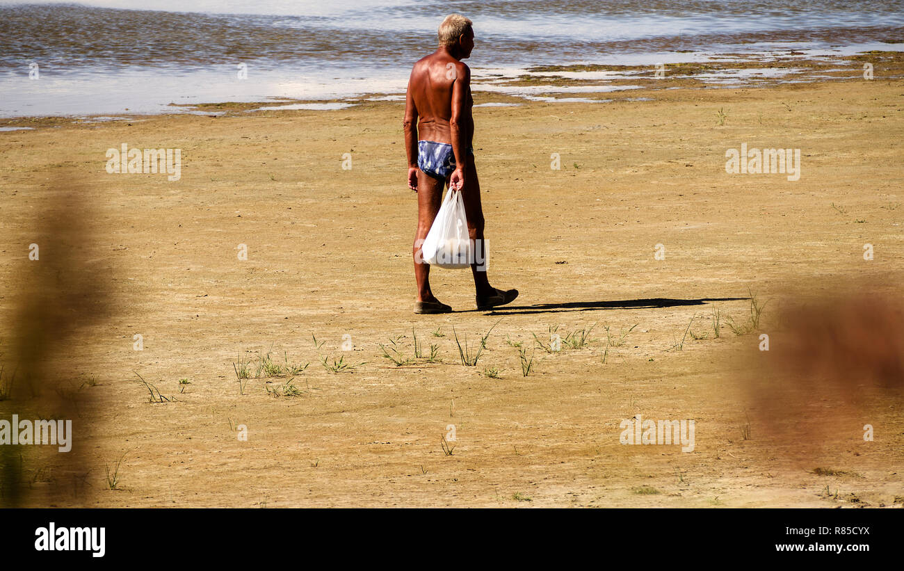 Single senior retired sun-tanned man walking on the beach looking at the ocean with plastic bag.  Copy space. Stock Photo