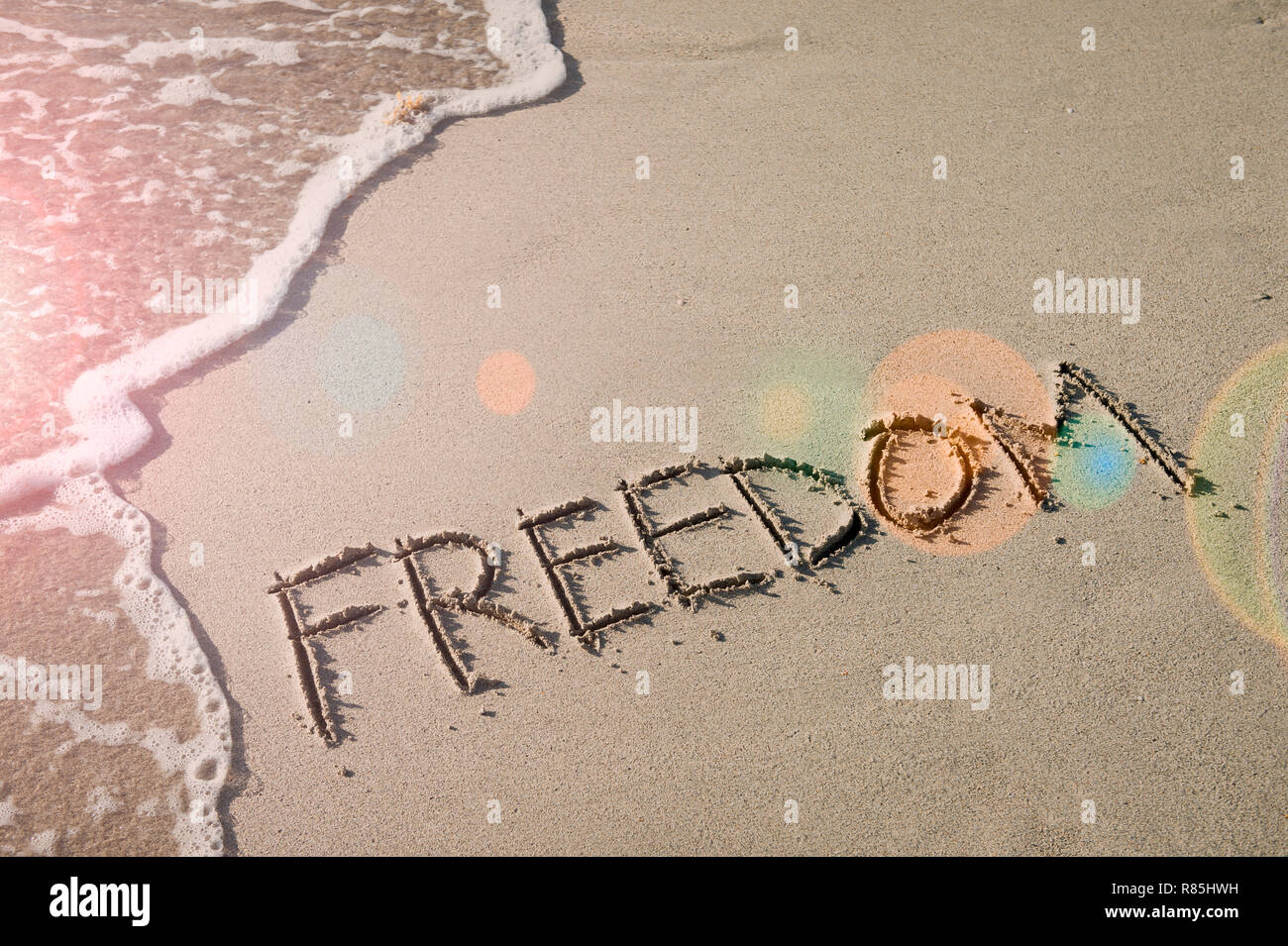 Freedom message handwritten in smooth sand with inspirational lens flare next to oncoming wave Stock Photo