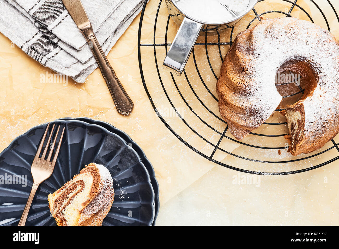 Marble bundt cake on french cooling rack with powdered sugar and one slice of cake on dark blue side plate over baking paper. Holiday baking backgroun Stock Photo