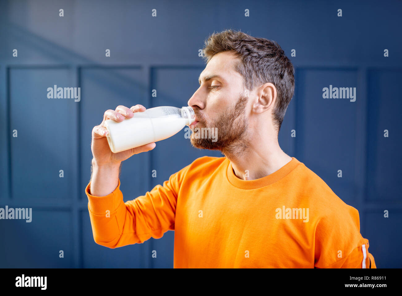 Conceptual portrait of a sports man in bright sweater drinking milk from the bottle on the blue background Stock Photo