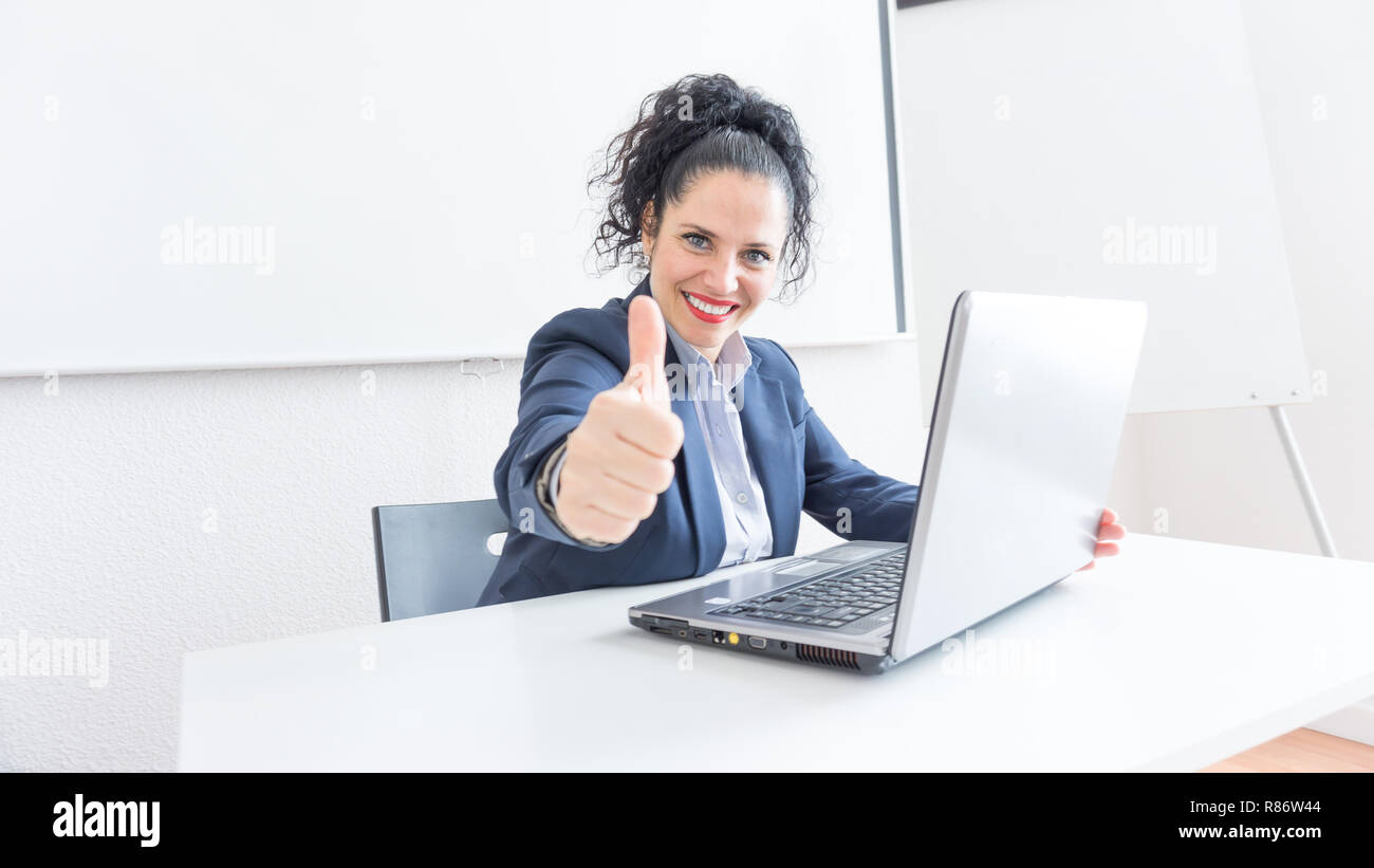 Portrait of a normal business woman doing thumb up sign in a office desk. With copy space. Dressing professionally smiling & looking at the camera. Black hair and blue eyes Stock Photo