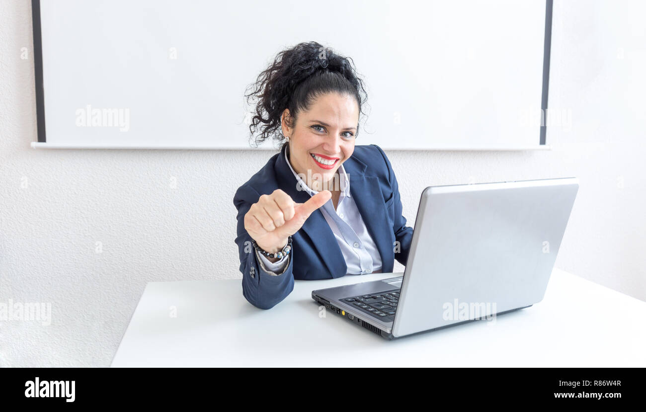 Portrait of a normal business woman doing thumb up sign in a office desk. With copy space. Dressing professionally smiling & looking at the camera. Black hair and blue eyes Stock Photo