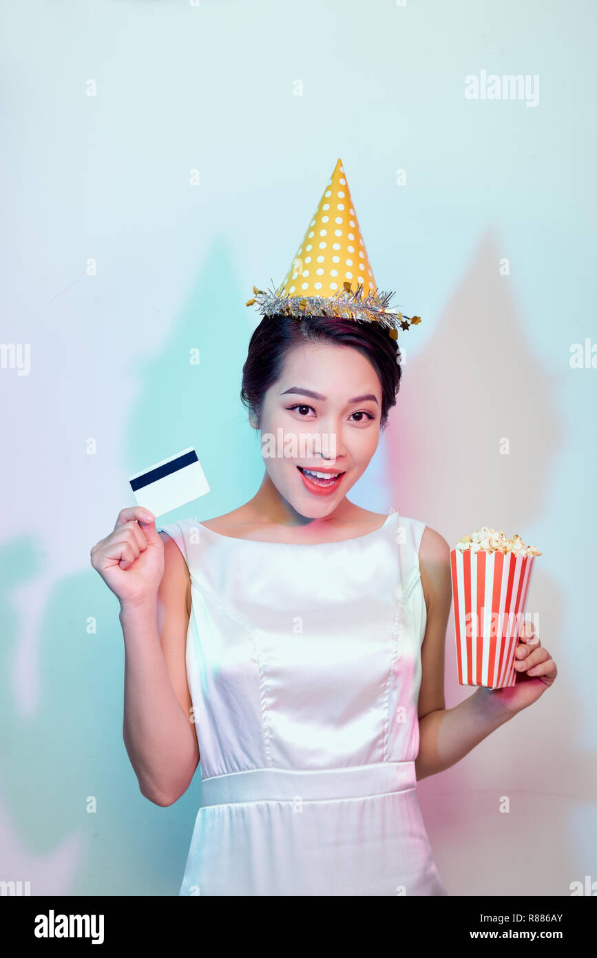 Portrait of young overjoyed attractive woman in white dress watching movie film, holding bucket of popcorn and credit card isolated on white backgroun Stock Photo