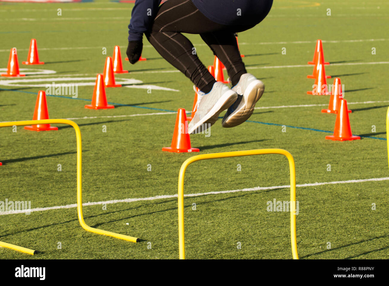 An athlete works out jumping over yellow hurdles on a green turf field with orange cones on it Stock Photo