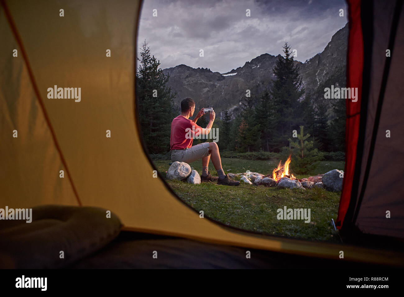 View from inside tent at sunset. Man hiker sitting at bonfire with cellphone Stock Photo