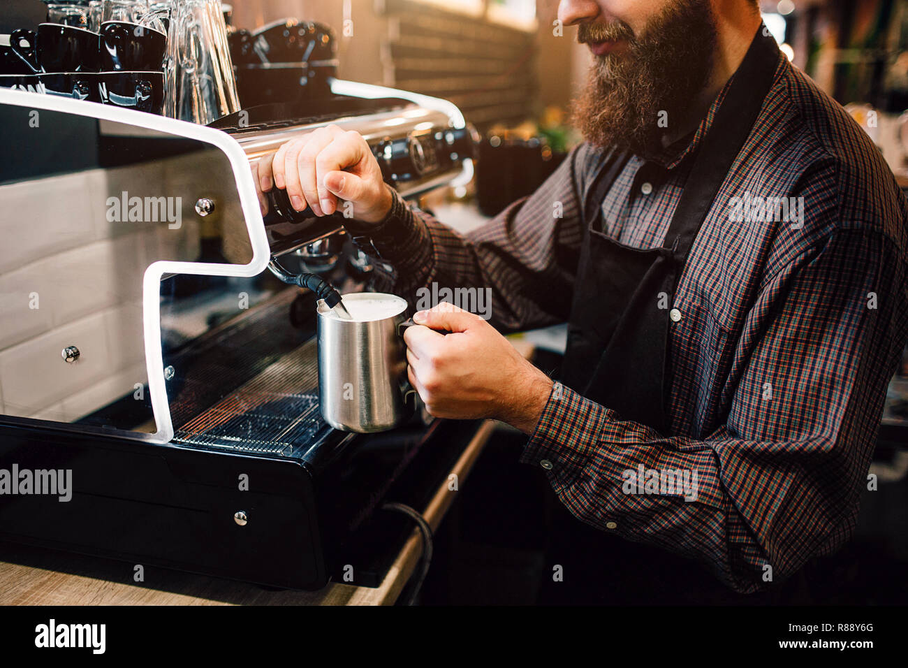Young bearded barista floating milk. He does it careful. Guy wears apron. Young man prepares milk for coffee Stock Photo