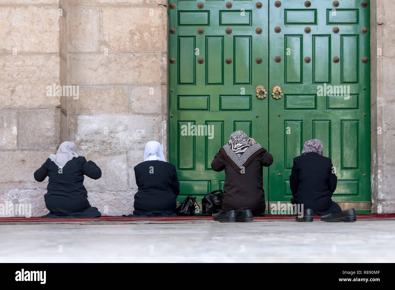 four women pray on their knees in front of the green closed door of the old mosque Dome of the Rock in the Muslim quarter of the old city of Jerusalem Stock Photo
