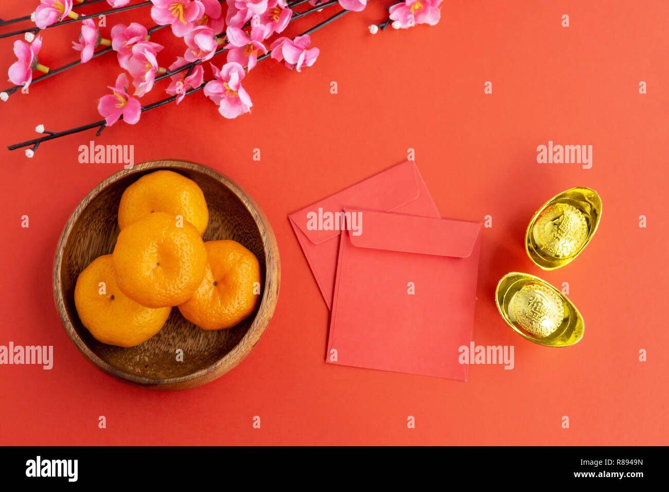 Flat lay Chinese New Year Background - Cherry Blossom, Mandarin Orange, Red Envelop and Golden Ingots on red background. Stock Photo