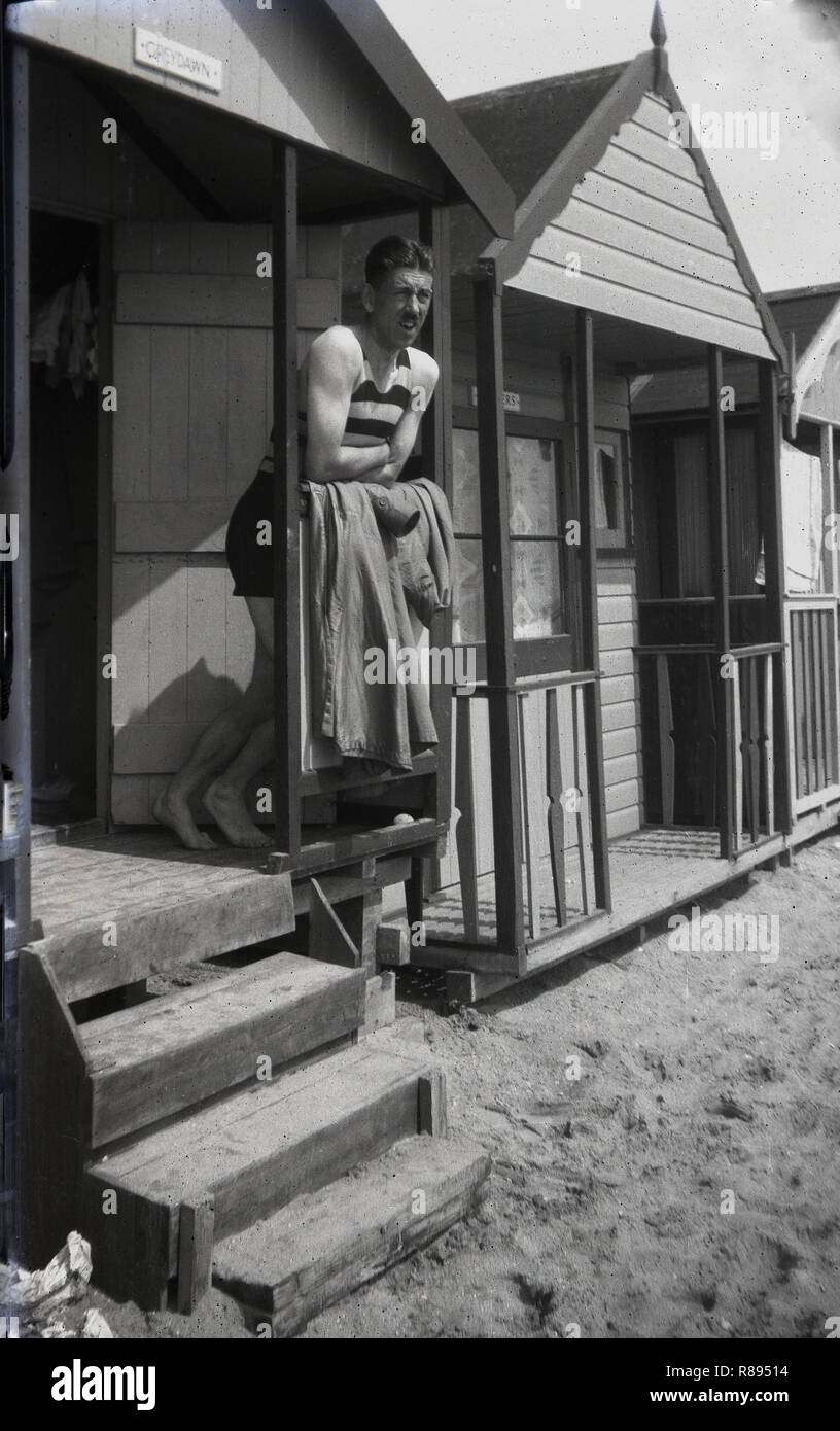 1930s, an English man in a stripy one-piece sleeveless swimsuit of the era, standing on the veranda of a beach hut at Southend-on-sea, England, a popular holiday resort for people living in Essex and easr London at this time. Stock Photo