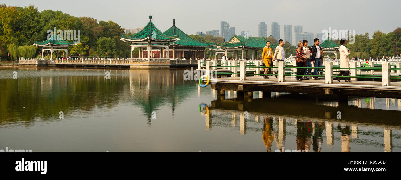People walking along elevated walkways over East Lake, in Wuhan, China. the green roofed pavilions on walkway are reflected in the water. Stock Photo