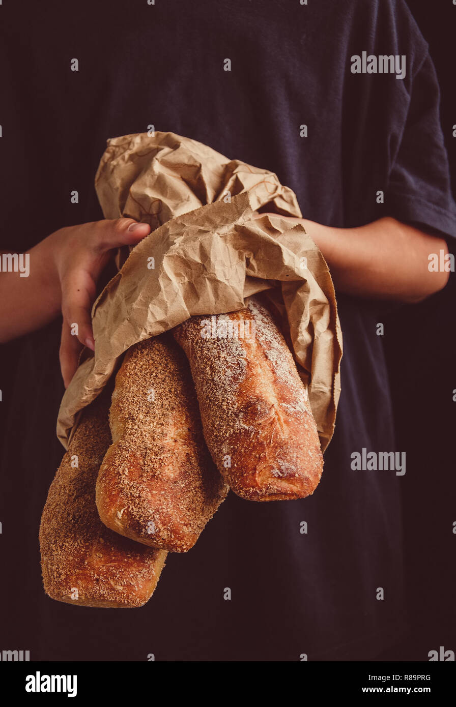 Freshly baked baguettes in the craft paper bag in the boy's hands. Dark food photo. Rustic style. Selective focus. Toned. Stock Photo