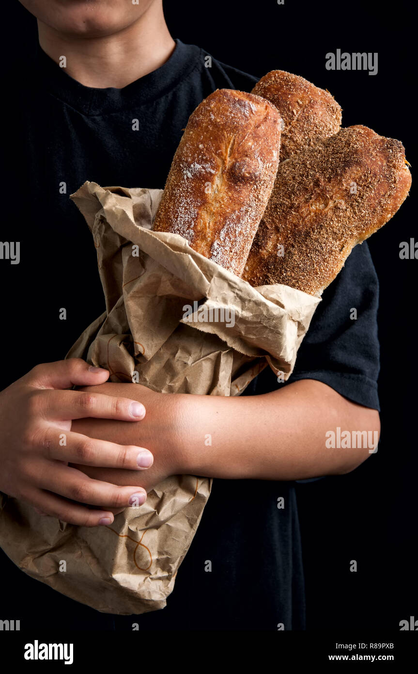 Freshly baked baguettes in the craft paper bag in the boy's hands. Dark food photo. Rustic style. Selective focus. Stock Photo
