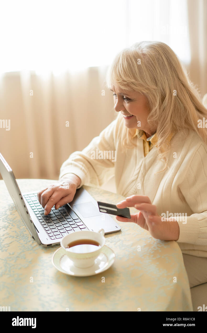 Beautiful Mature Woman Makes Purchases On The Internet With A Credit Card While Sitting At The Table And Having A Cup Of Tea Stock Photo