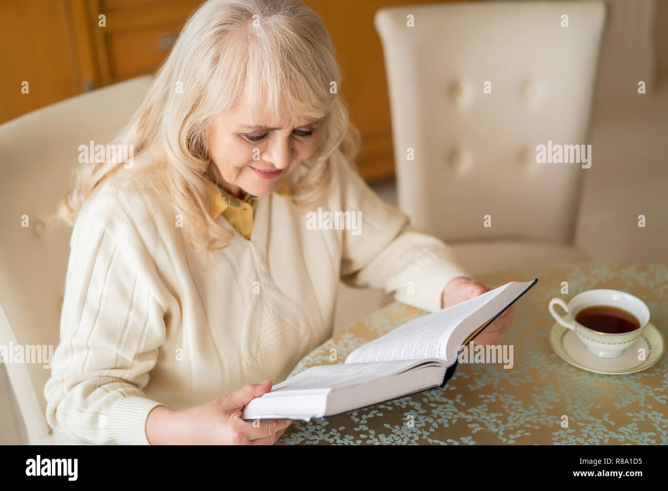 Concentrated Grandmother Reads A Book At The Table While Having A Cup Of Tea In The Living Room Stock Photo