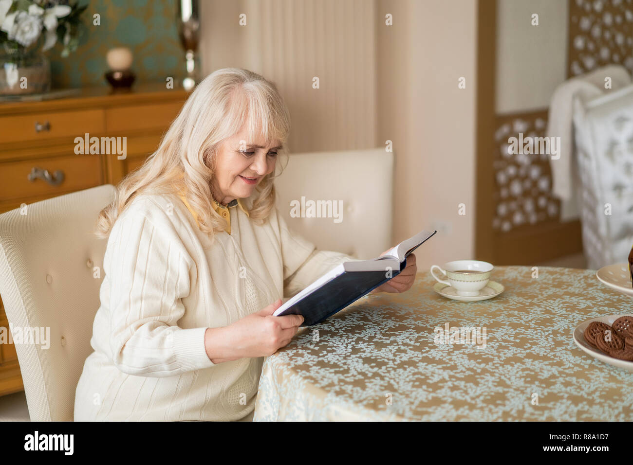 Horizontal Shot Of Beautiful Elderly Woman Over The Book At The Table While Having Tea And Cookies. Stock Photo