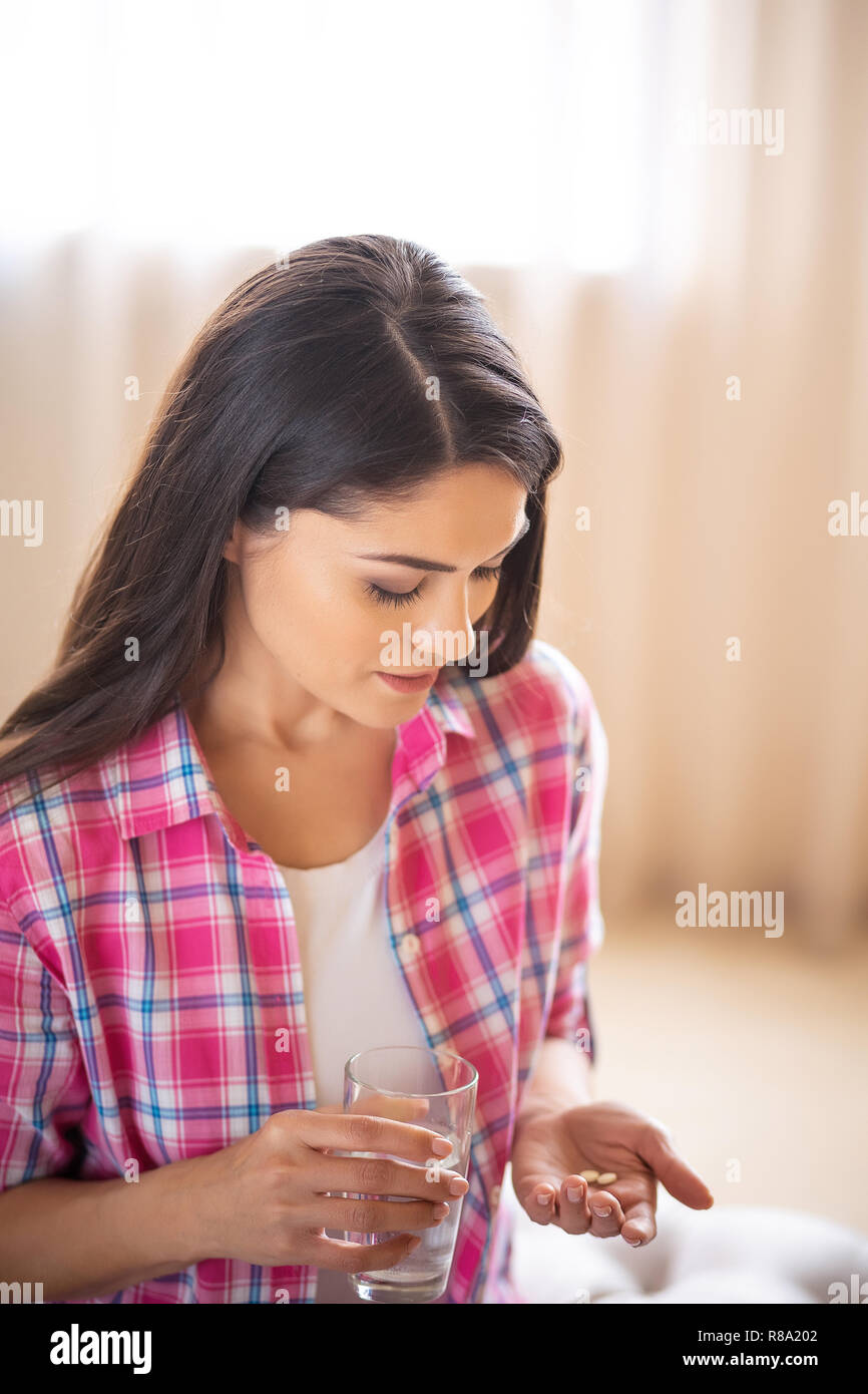 Brunette Girl Pensively Looks On The Tablets From Headache In Her Hand And Holds A Glass Of Water While Sitting On The Sofa. Stock Photo
