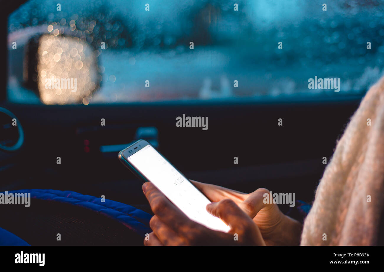 A woman sits in the passenger seat of a car and uses a smartphone in the early winter morning. Stock Photo