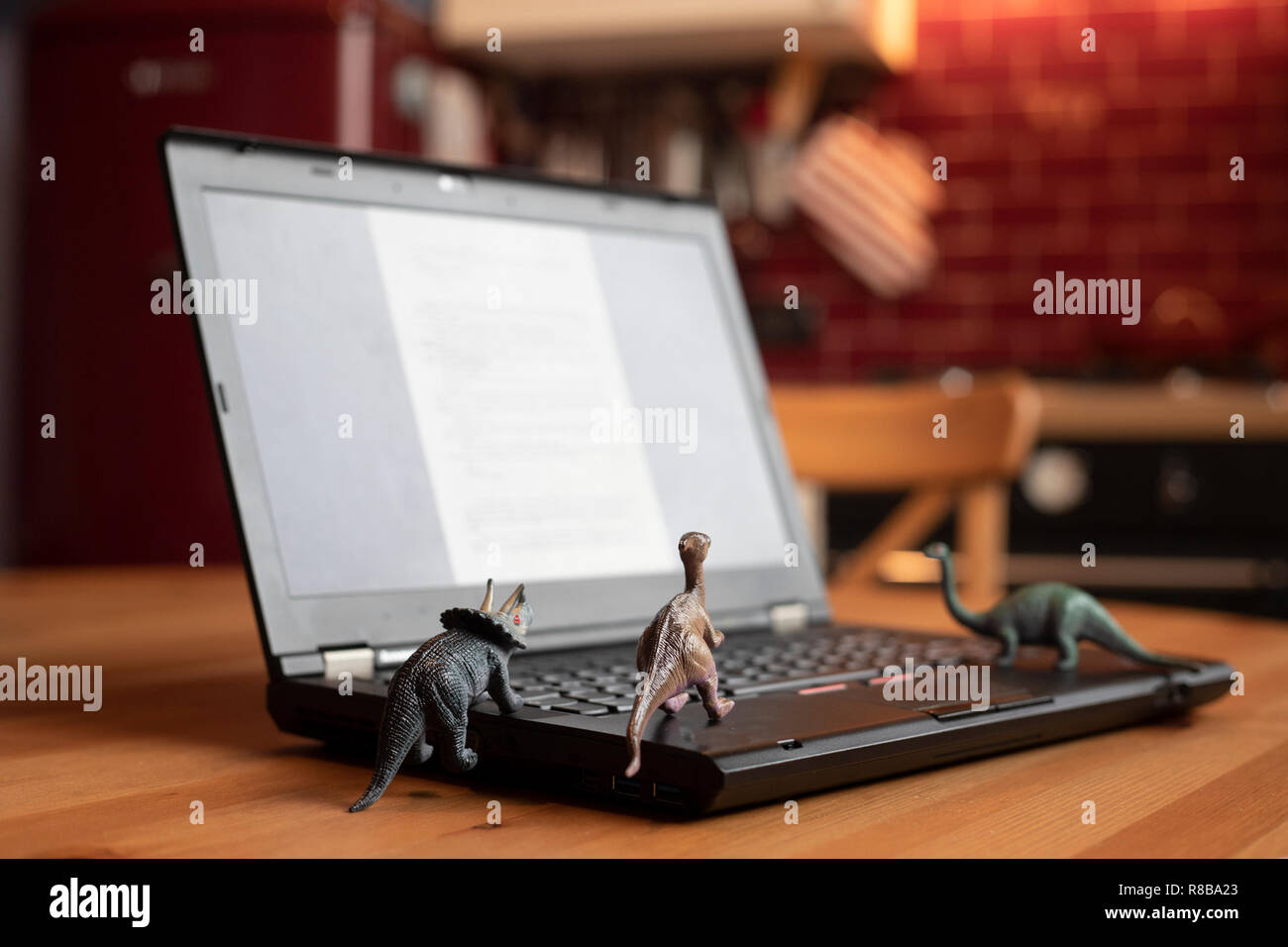 Open laptop on wooden table with small dinosaur toys that seem looking at the screen. Natural light. Smartworking during lockdown symbol. Stock Photo