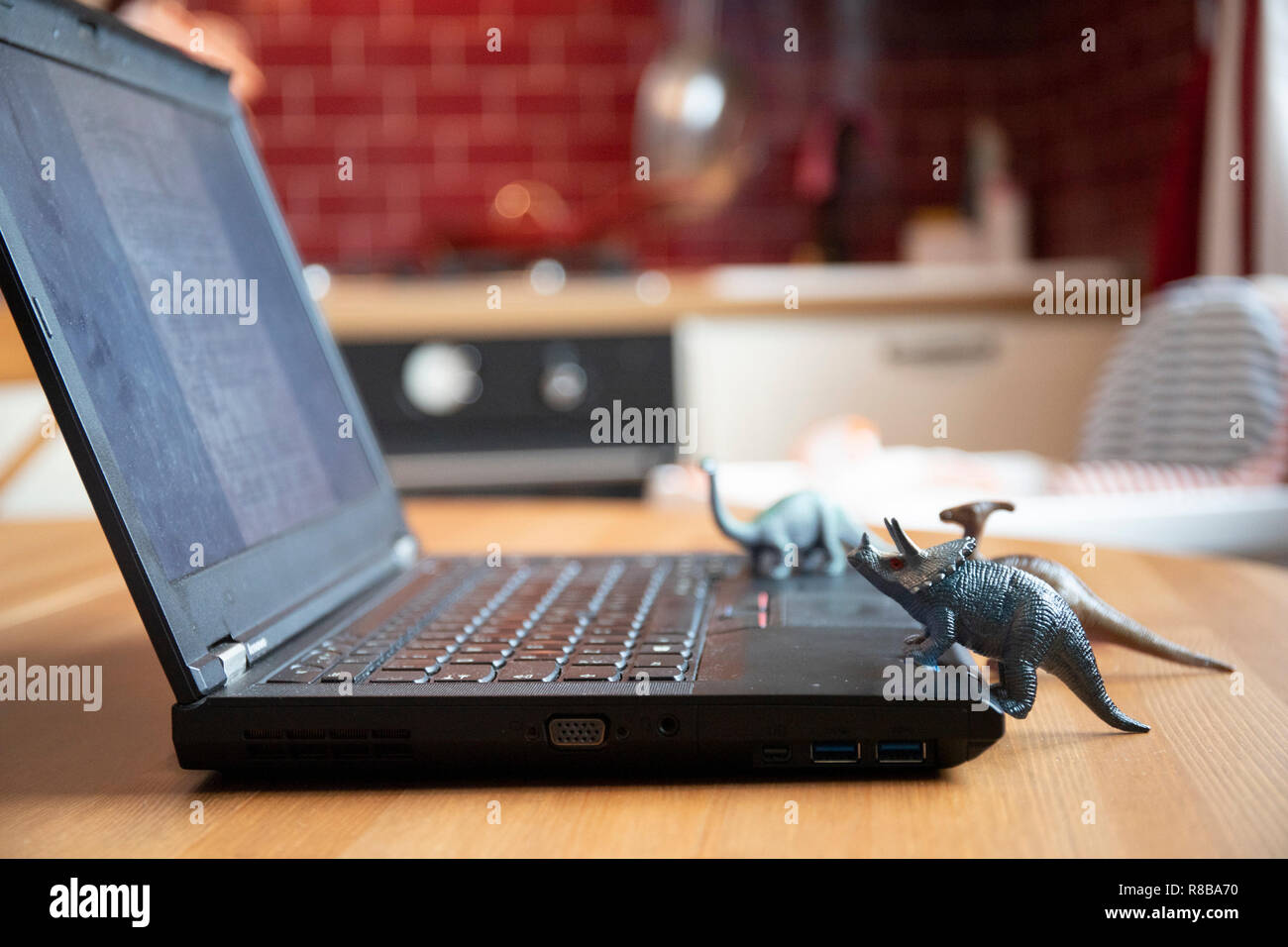 Open laptop on wooden table with small dinosaur toys that seem looking at the screen. Natural light. Side view. Stock Photo