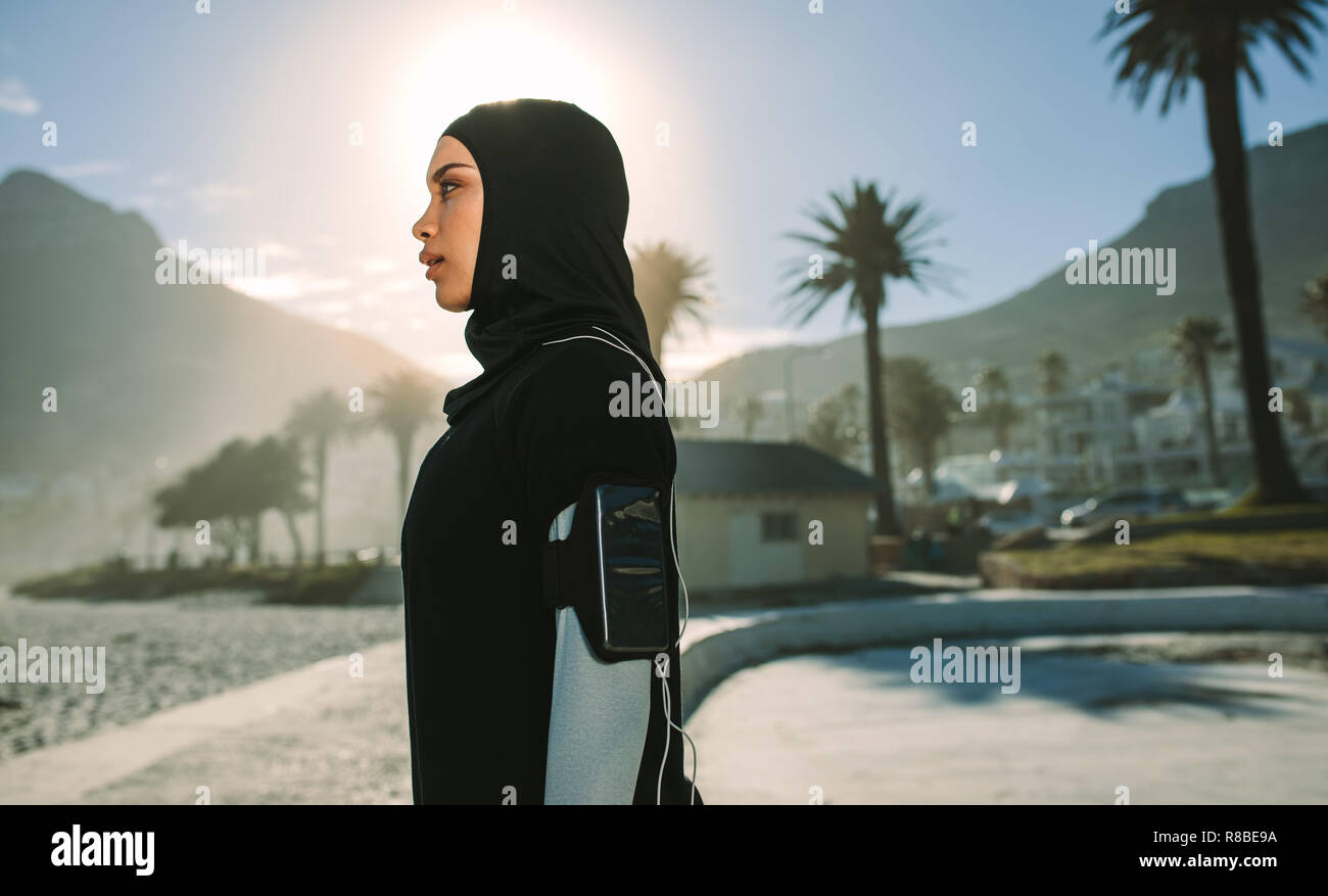 Side view of young hijab woman standing outdoors with bright sunlight from back. Muslim woman resting after a morning run in the city. Stock Photo