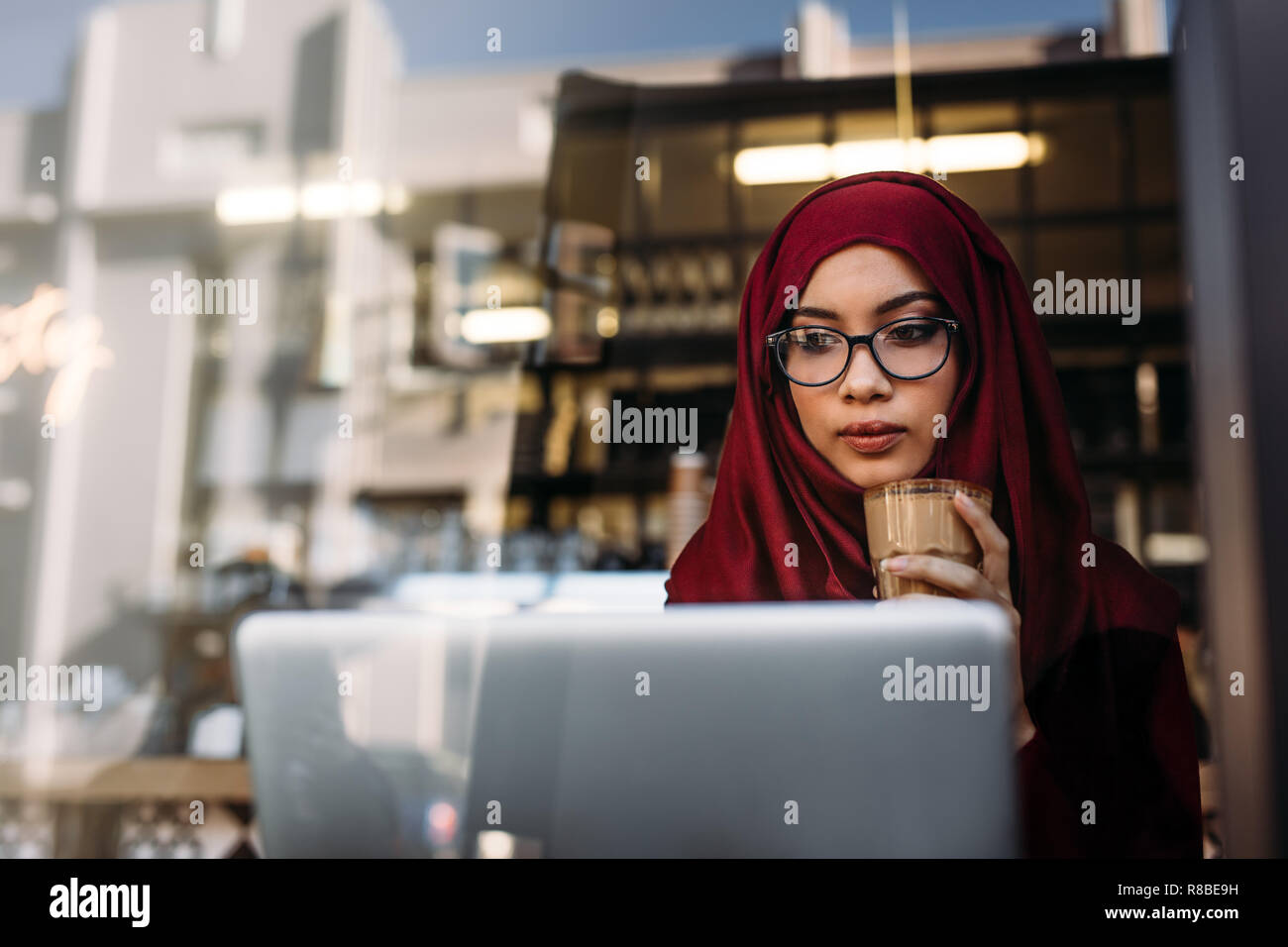 Young woman wearing hijab and eyeglasses using laptop with coffee in hand while sitting at cafe. Muslim female having coffee at cafe and using laptop Stock Photo
