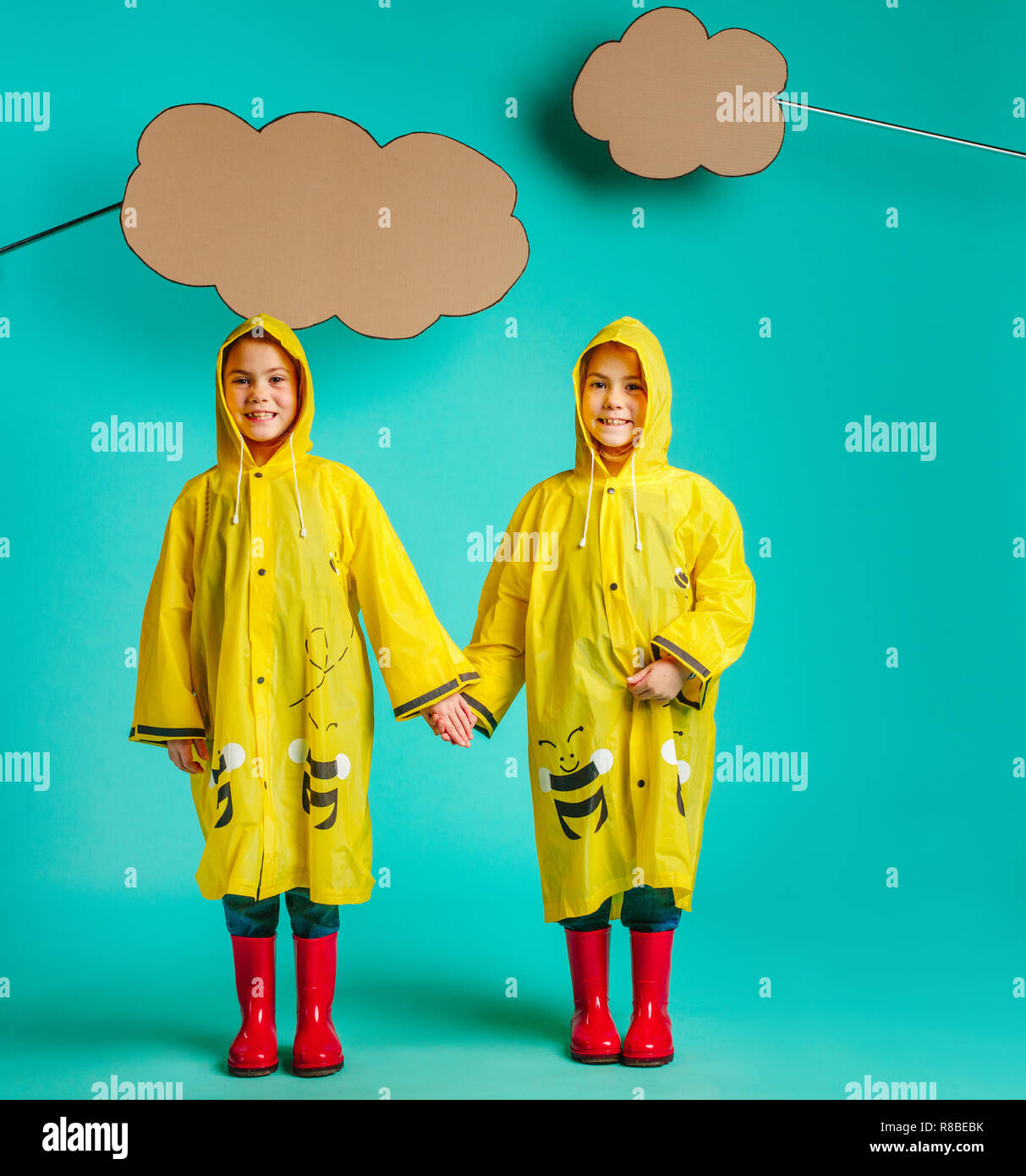 Happy twins girls wearing raincoat standing together holding hands under paper clouds. Small girls in rainwear standing on blue background. Stock Photo