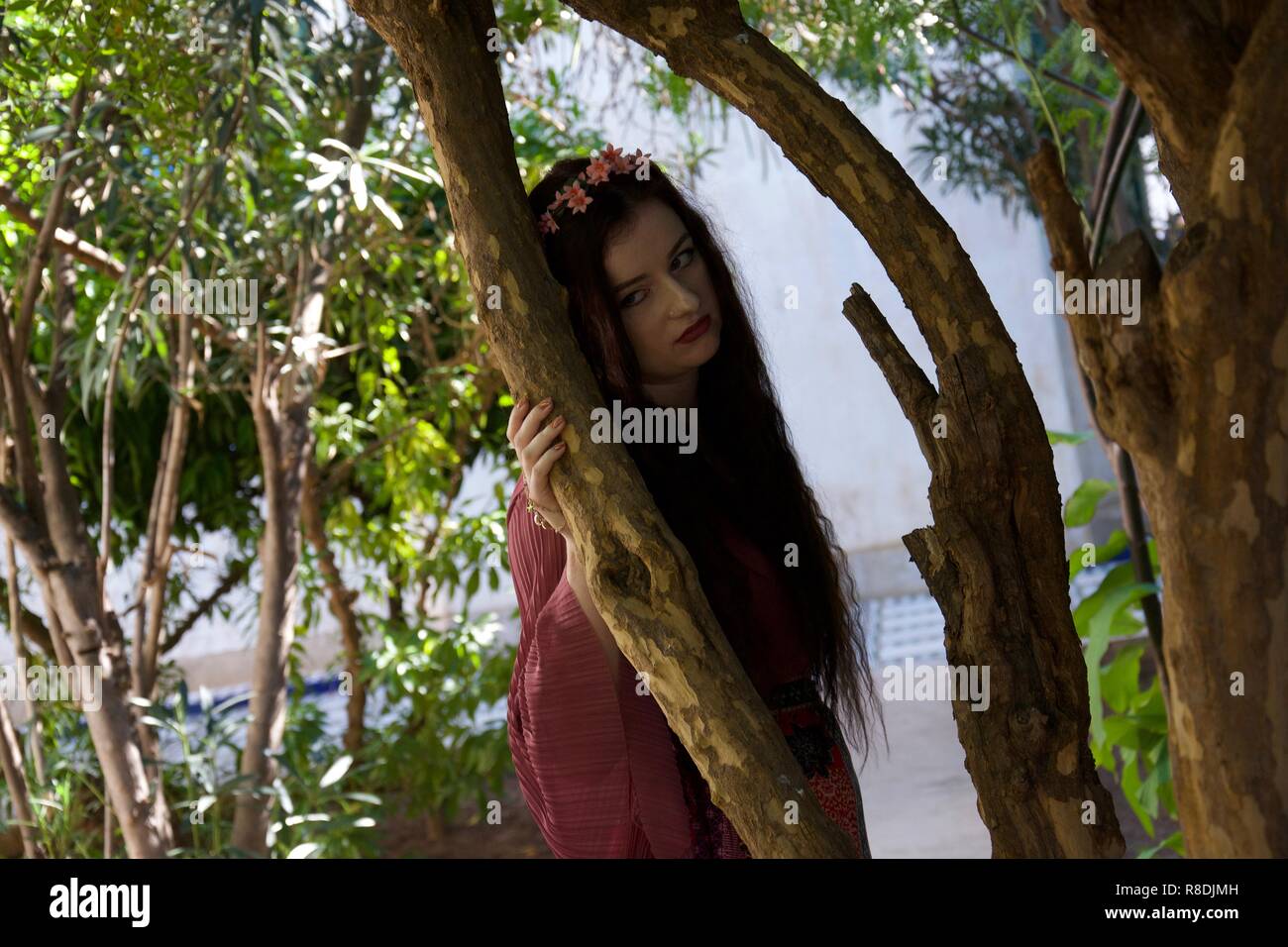 A hippie girl with flowers in her long hair leans against a tree, sulking and looking petulant and fed up Stock Photo