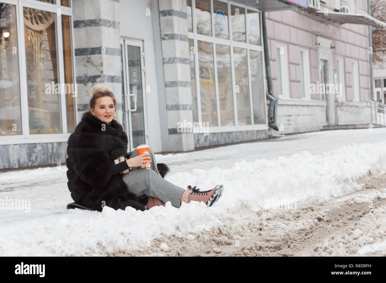girl sitting on a snow in the city on curb covered with snow. Lifestyle. winter mood, coffee cup, mink fur clothing. Stock Photo