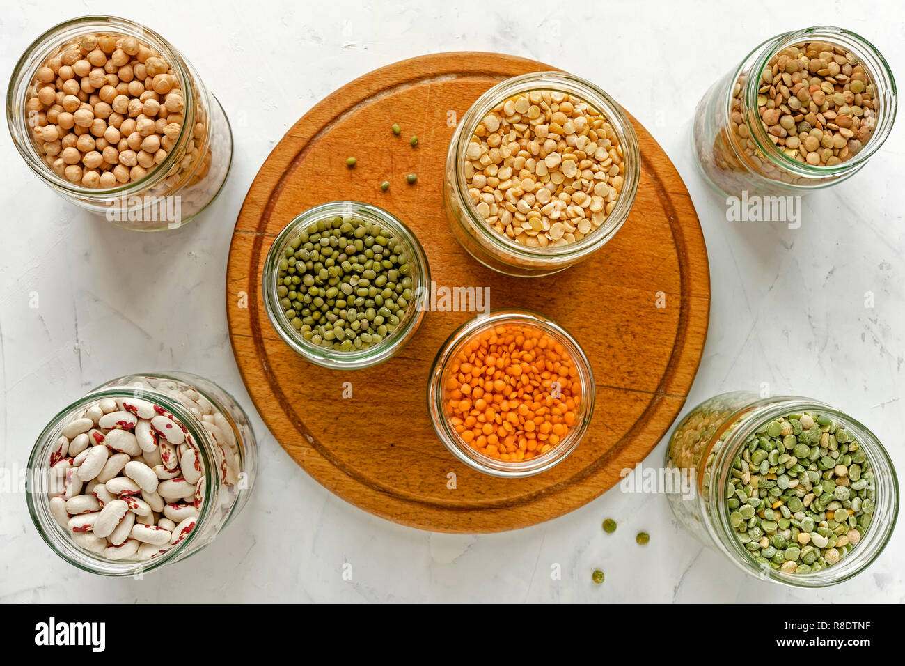 Assorted dry legumes and pulses in glass jars around a circular wooden board on white viewed from overhead in a healthy diet concept Stock Photo