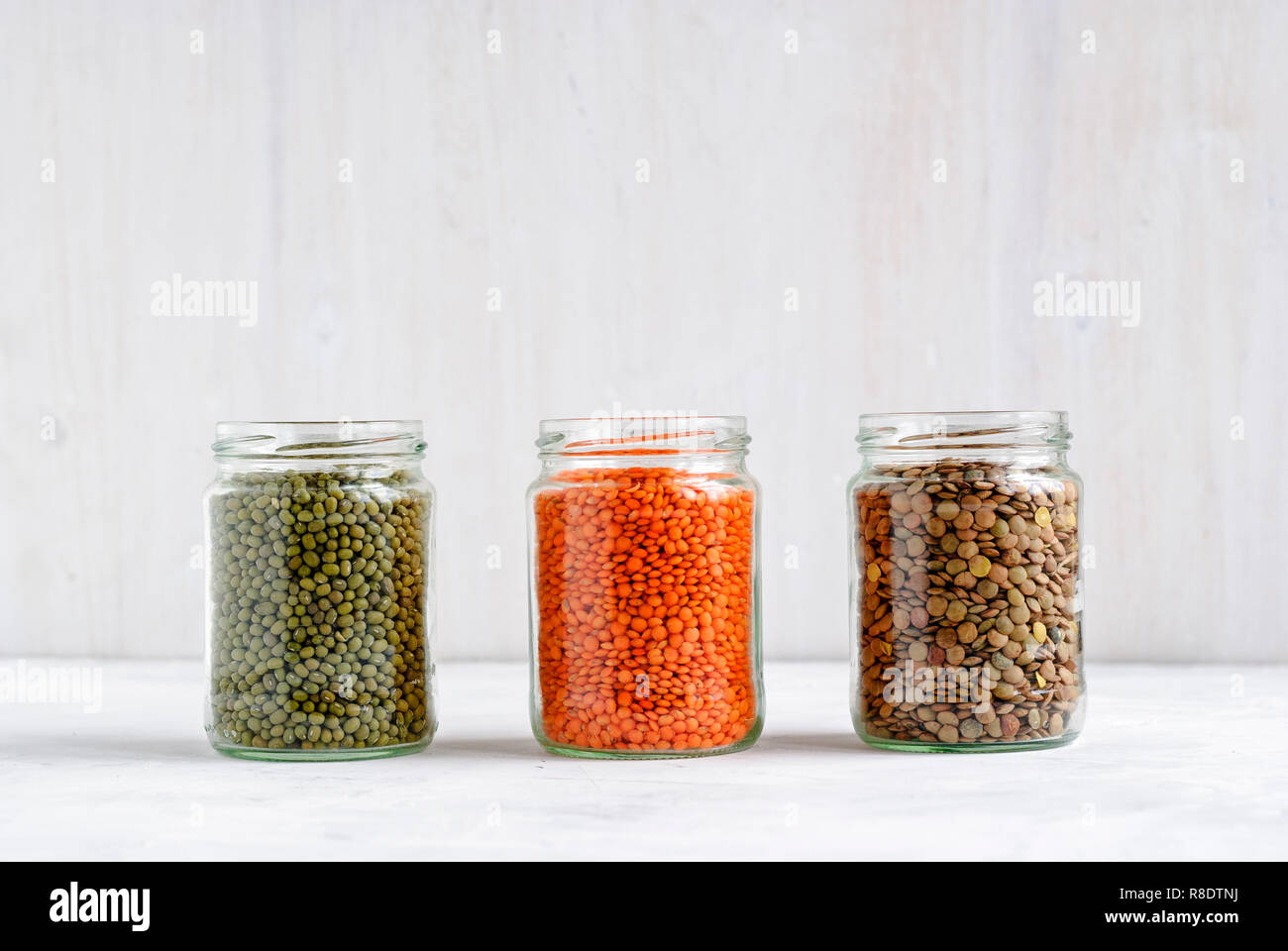 Dried Mung beans, and green and red lentils in glass storage jars standing in a row on white for