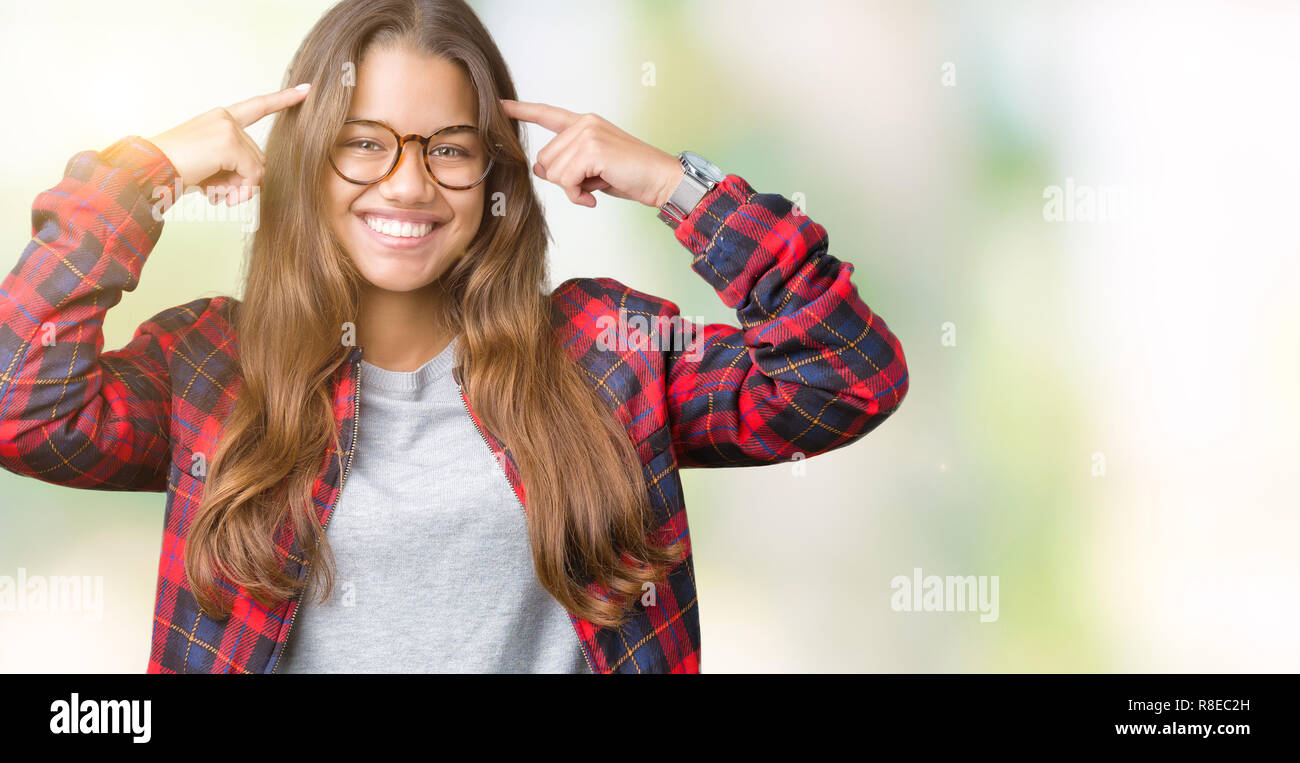 Young beautiful brunette woman wearing jacket and glasses over isolated background Smiling pointing to head with both hands finger, great idea or thou Stock Photo