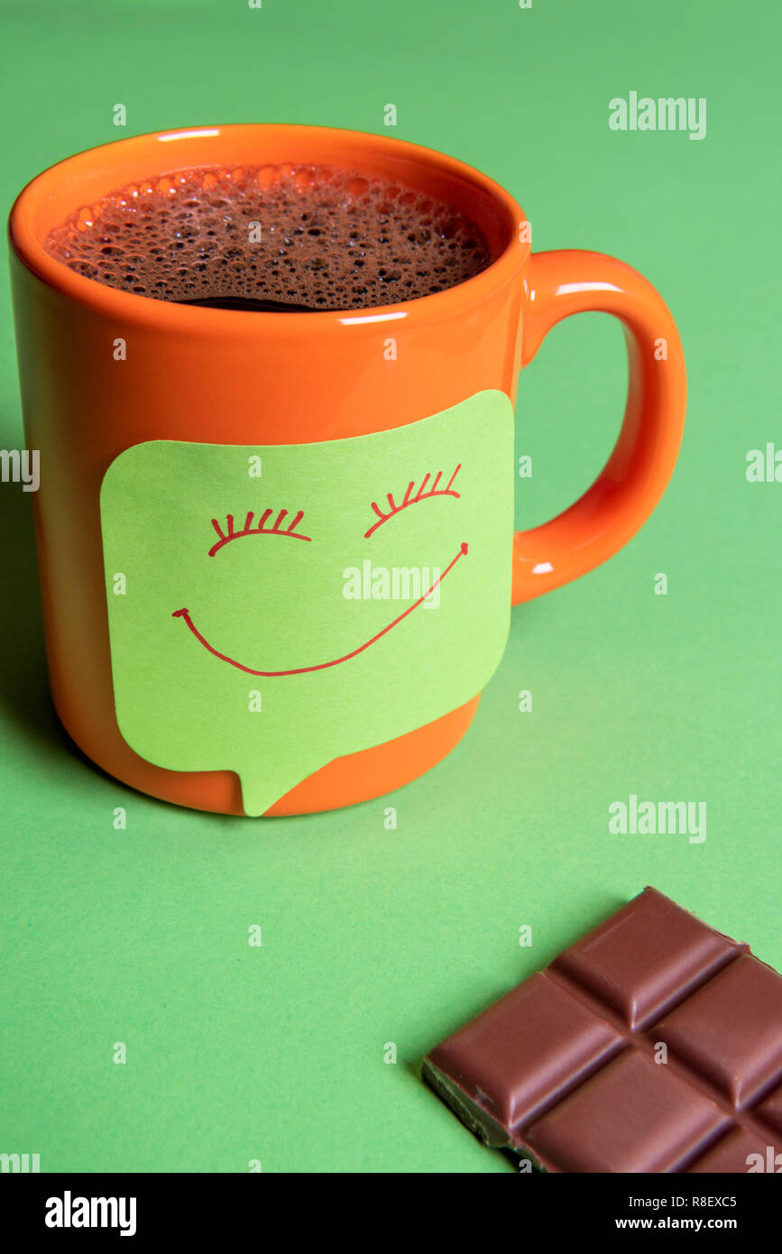 Fresh hot coffee in an orange mug and a funny smiley face sticky note on a green table beside a big piece of chocolate Stock Photo