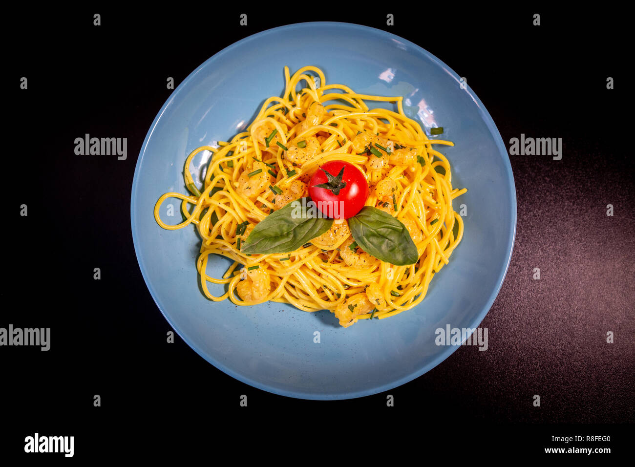 Top view of yellow spaghetti with prawns, a cherry tomato and basil in a blue plate on a black table. Stock Photo