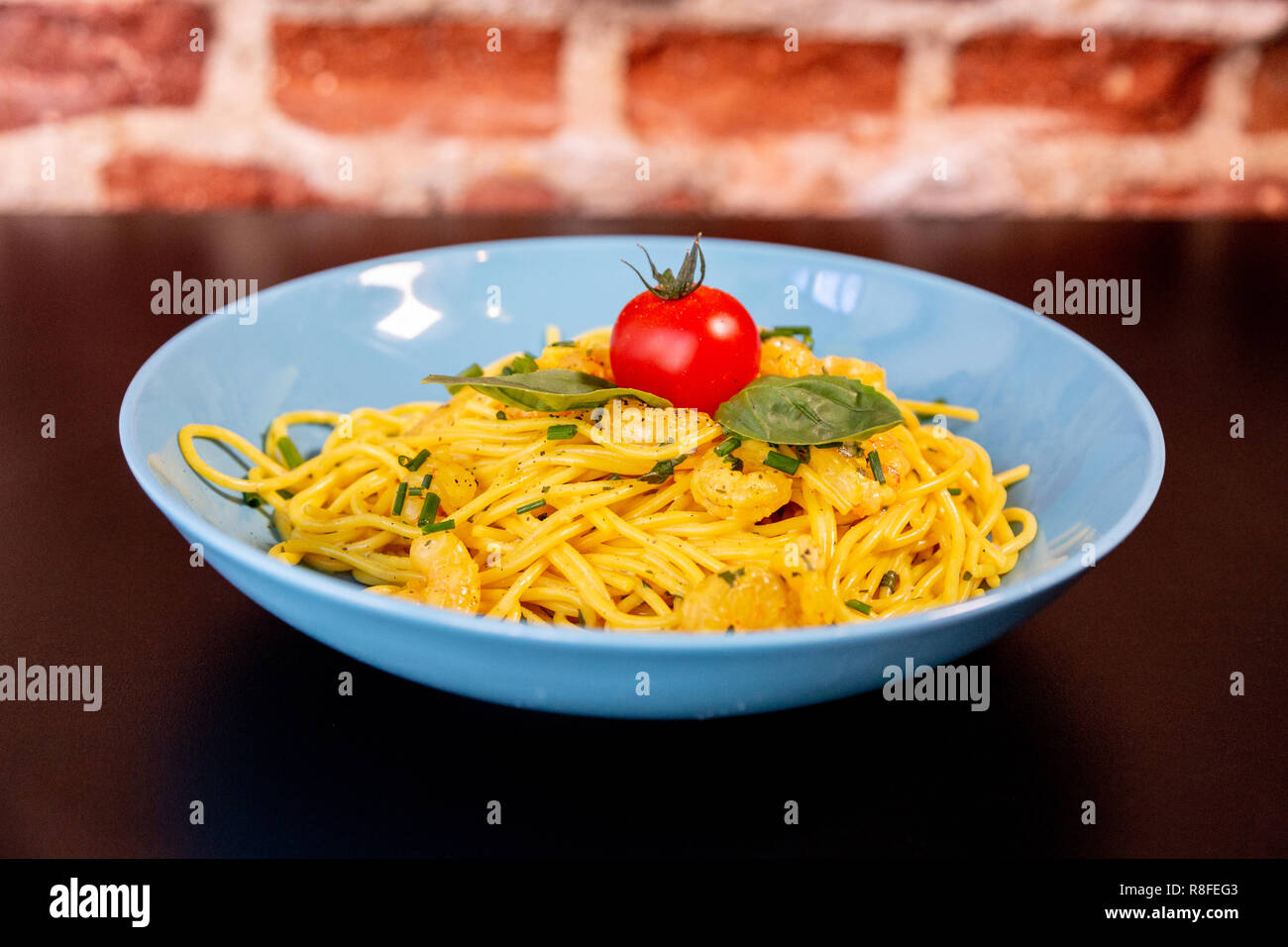 Yellow spaghetti with prawns, a cherry tomato and basil in a blue plate on a black table. With a brick wall. Stock Photo