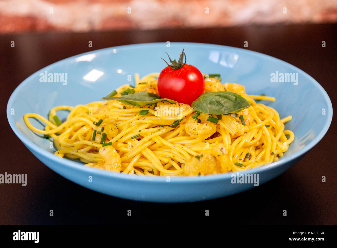 Close up of yellow spaghetti with prawns, a cherry tomato and basil in a blue plate on a black table. With a brick wall. Stock Photo