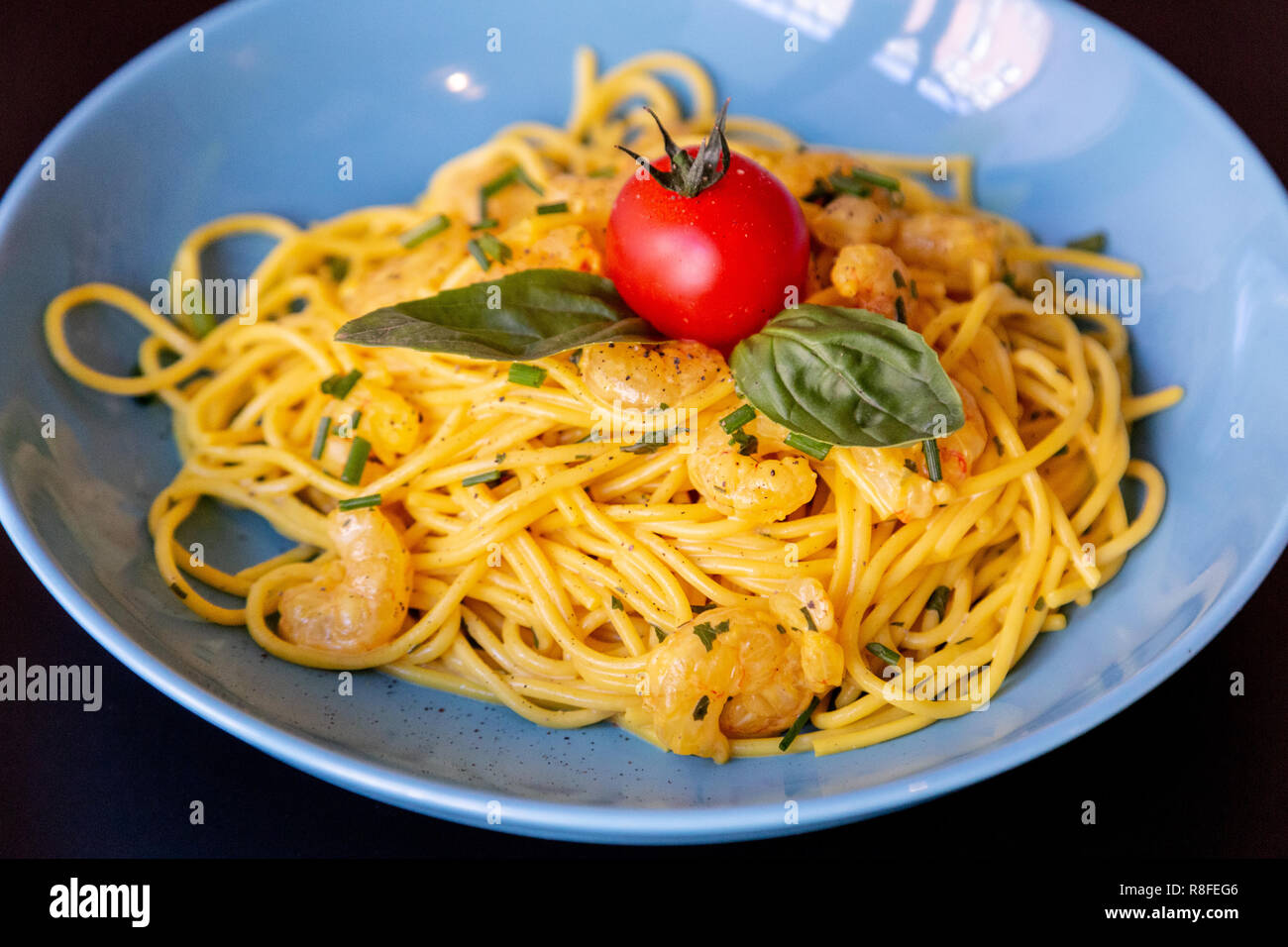 Close up of yellow spaghetti with prawns, a cherry tomato and basil in a blue plate on a black table. With a brick wall. Stock Photo
