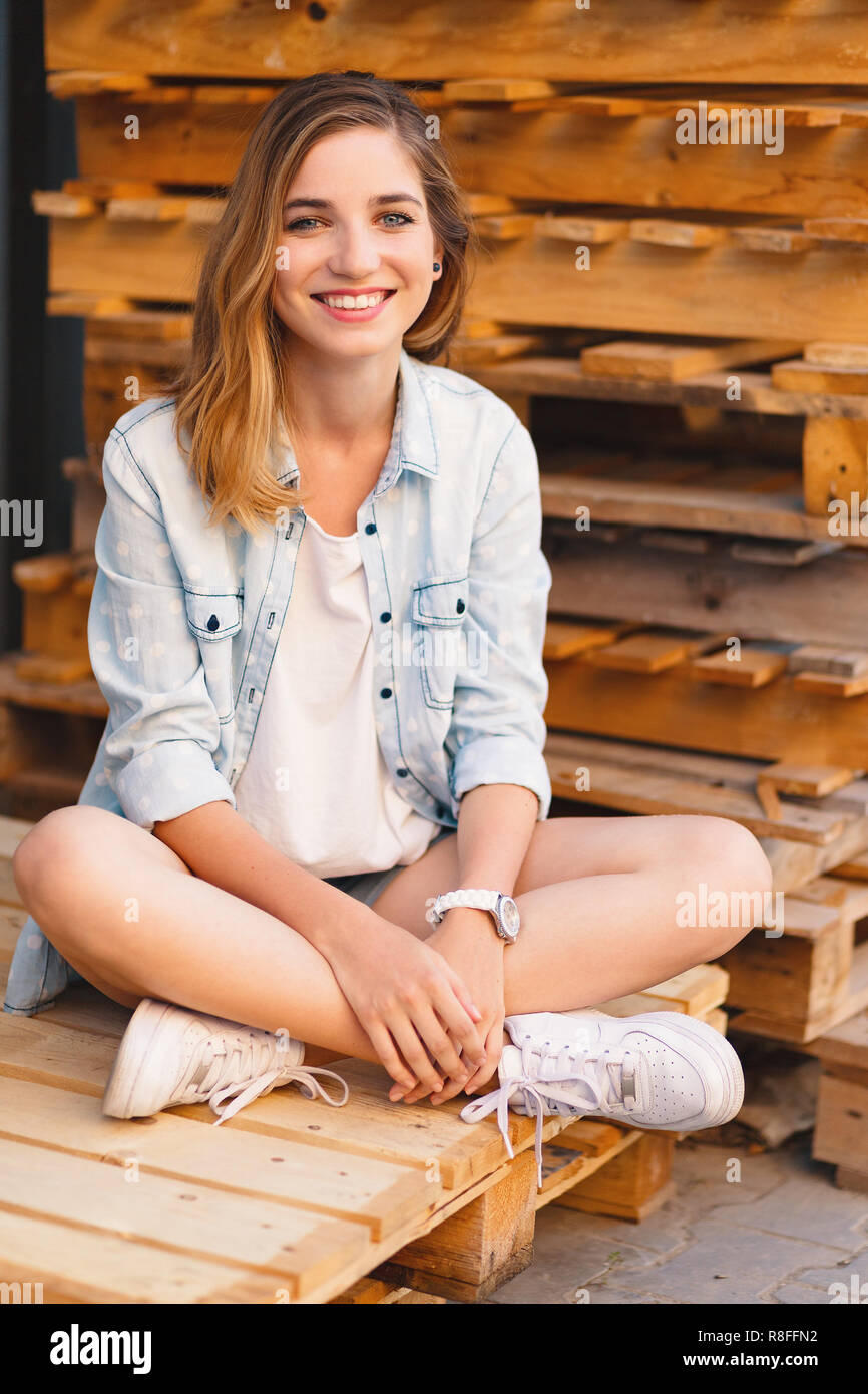 Pretty smiling girl, wearing jeans, shorts and shirt posing on wood pallet background during a sunny day. Stock Photo
