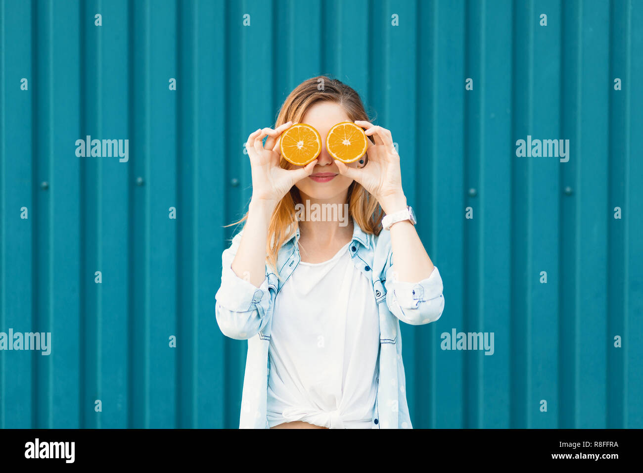 Carefree young beautiful girl using two halfs of oranges instead of glasses over her eyes over blue background. Stock Photo