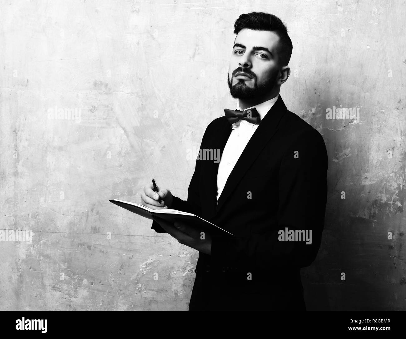 Proud and assured young employer with beard and in expensive suit writes plans in his organizer, on painted beige vintage background. Concept of confidence and business schedule Stock Photo