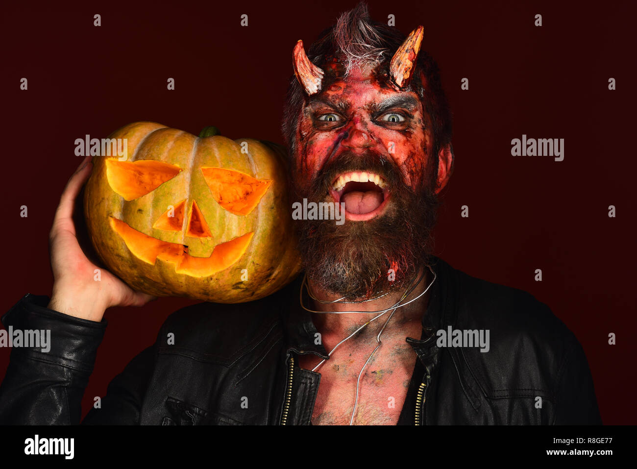 Demon with horns and evil smile holds carved jack o lantern. Man wearing  scary makeup holds
