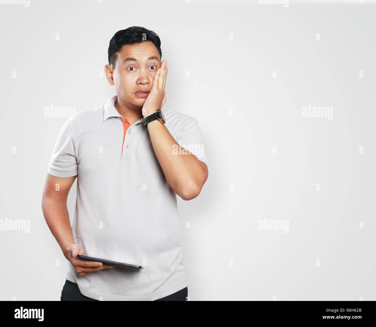 Photo image portrait of a cute handsome young Asian man with funny face looking shocked, closing his cheek with left hand while right hand holding tab Stock Photo