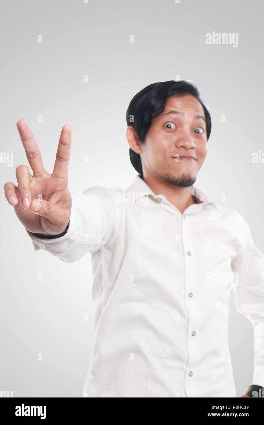 Photo image portrait of a cute funny young Asian man showing peace sign with smiling face, close up portrait over white background, focus on hand with Stock Photo
