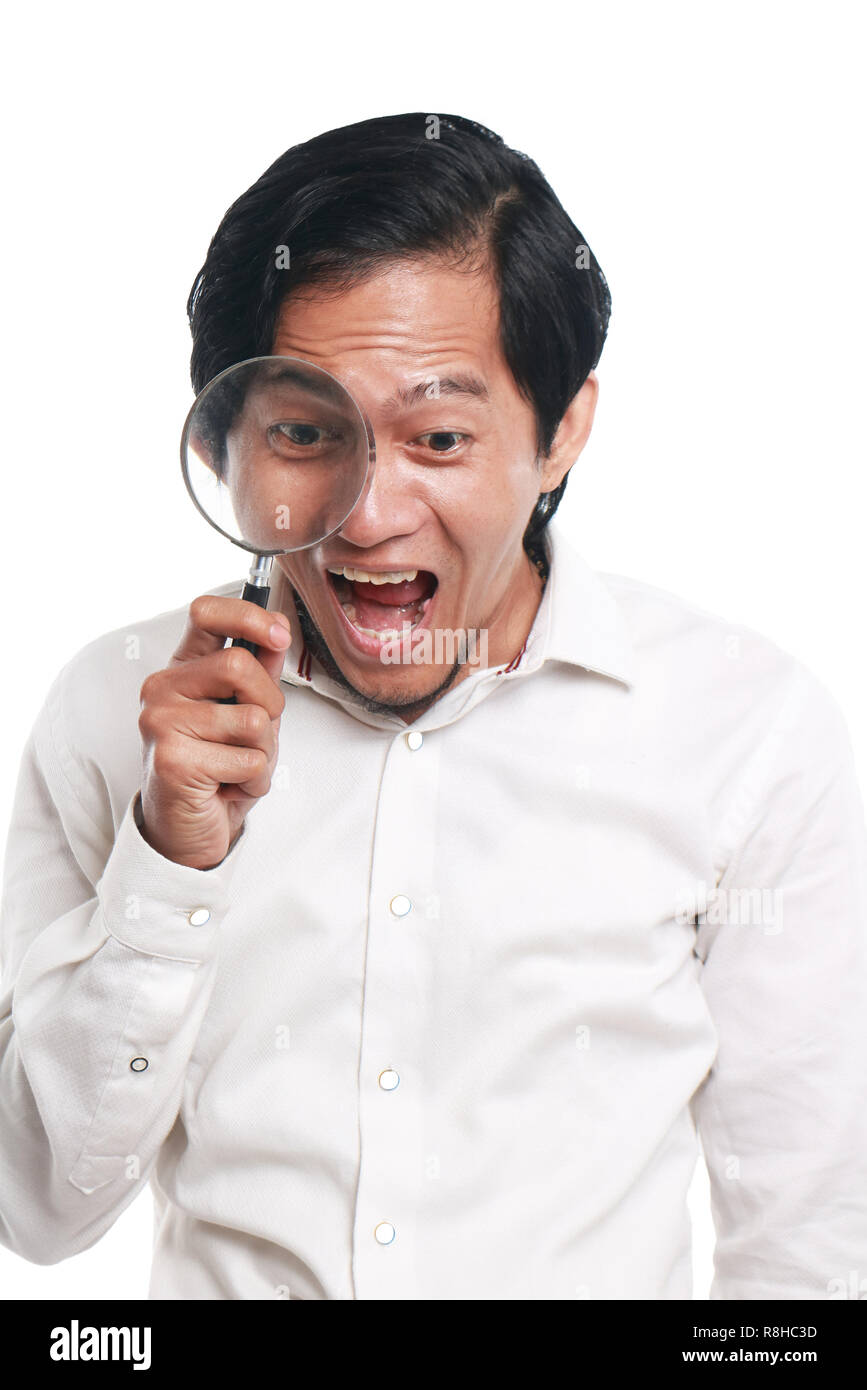 Photo image portrait of a funny young Asian businessman looked very happy, surprised and smiling to find something while looking into magnifying glass Stock Photo