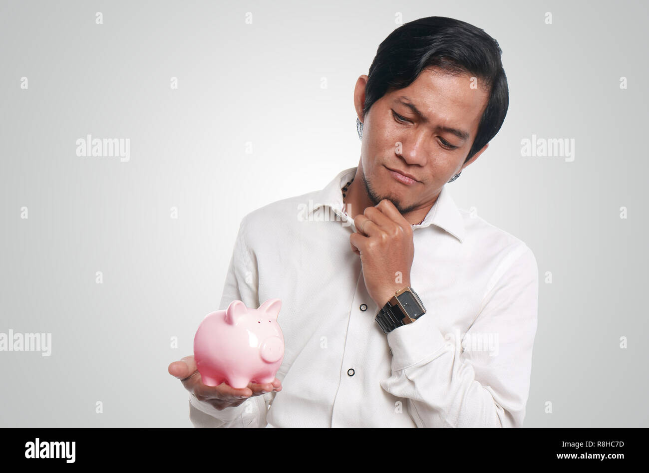 Photo image portrait of a funny young Asian businessman thinking something while looking a piggy bank on his hand, close up portrait, finance concept Stock Photo